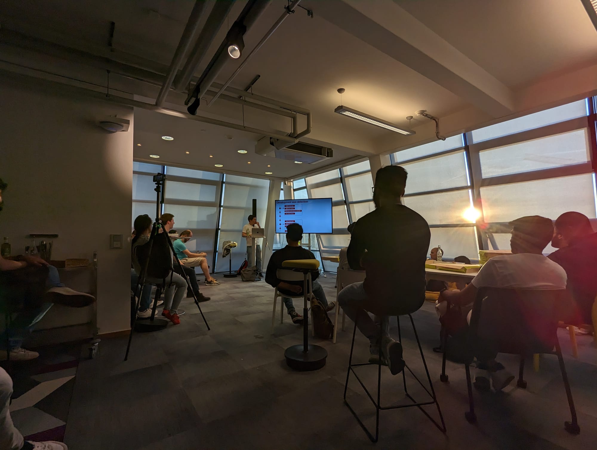 A wide-angle photo of a room of people listening to a speaker in front of a TV presenting something. On the left hand side near the wall, there is a tripod and a camera mounted on it, with a black backpack being used as weight for the tripod.